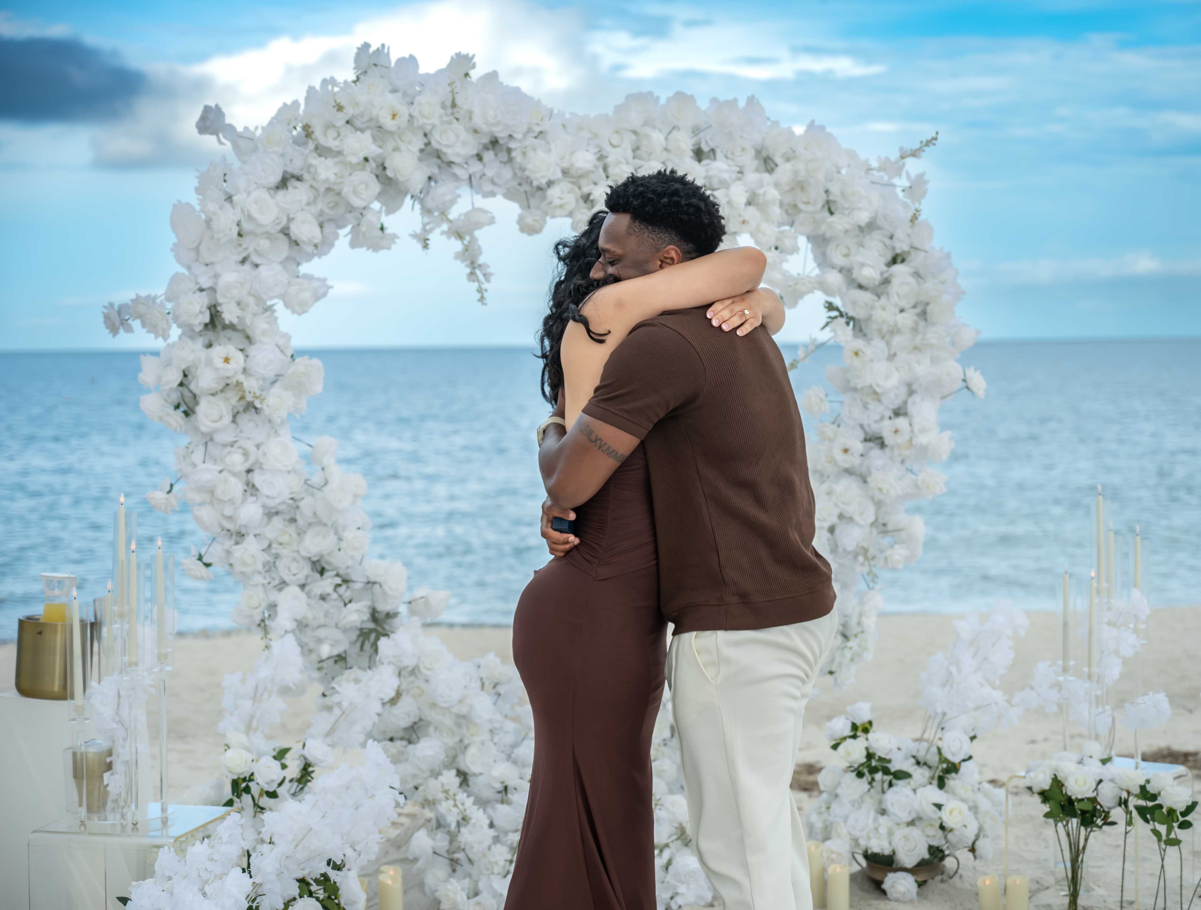 Couple embracing after a beach proposal, floral arch backdrop