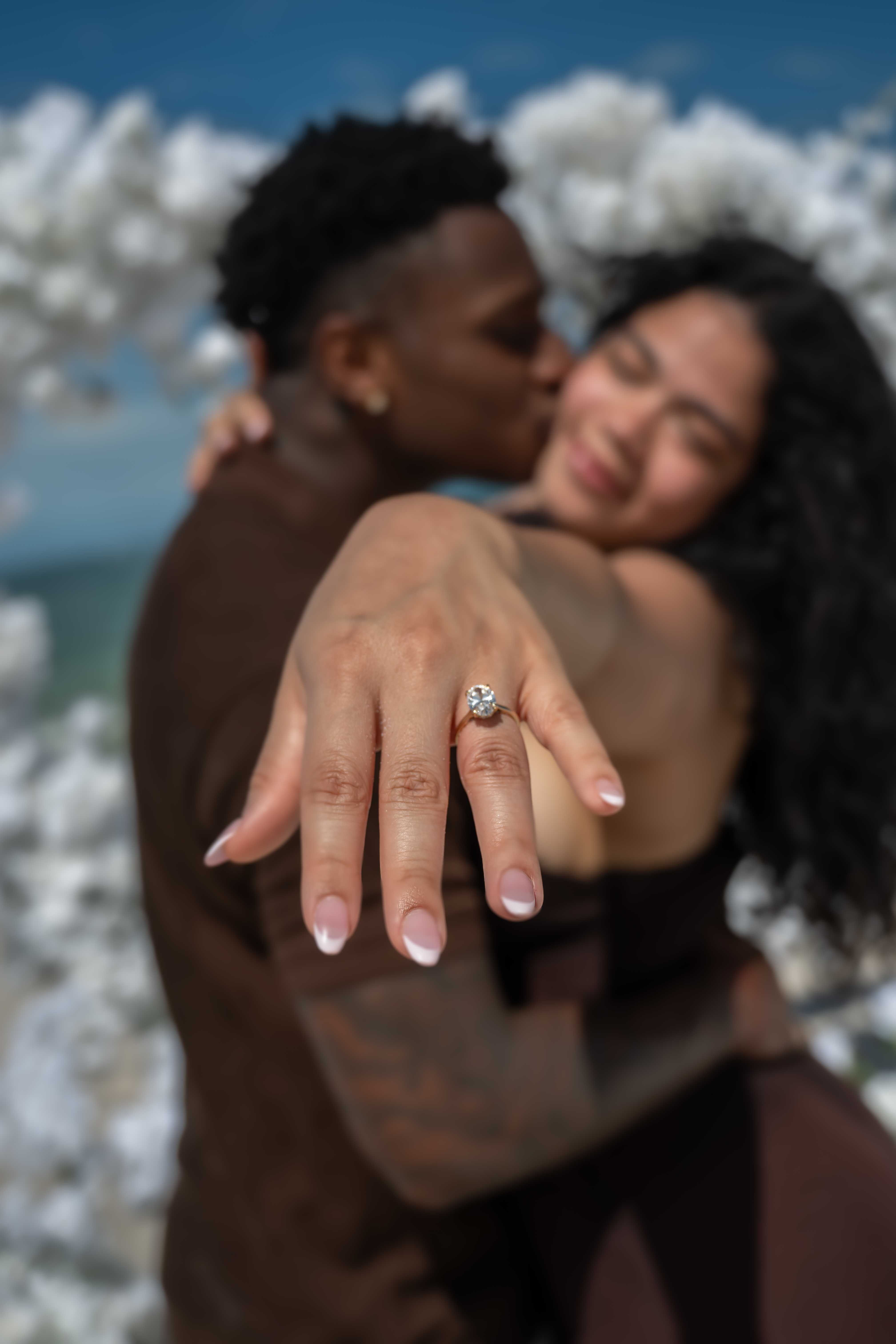 Engagement ring detail with the couple kissing in the background